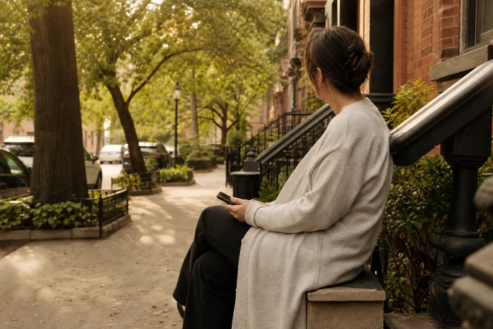 Woman sitting on a front stoop with a phone in her lap, green street trees
