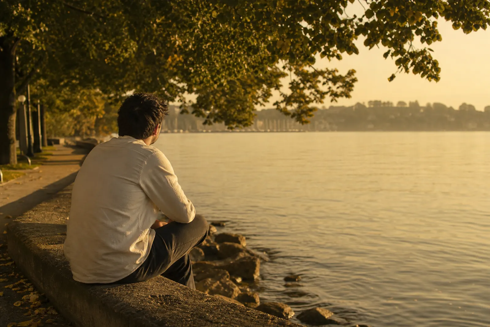 Man sitting on a low seawall at a waterfront park at golden hour, looking over calm water