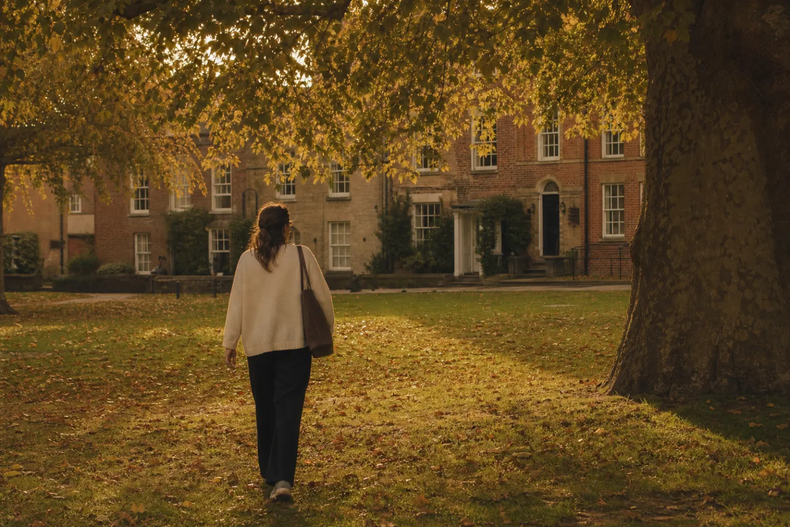 Woman crossing a leafy town green toward old brick and stone buildings in autumn