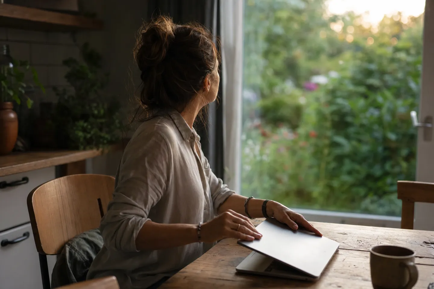 Woman pausing at a table with a closed laptop and looking into a garden